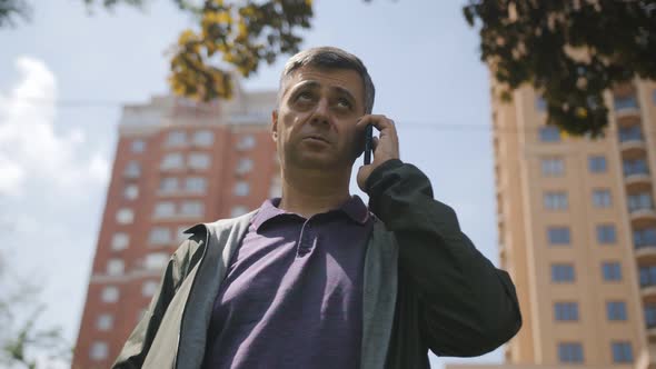 A Smiling Middleaged Man in the City Center Talks on the Phone Against the Background of Highrise