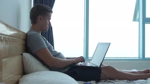 Young Man Sitting on Bed Using His Laptop in the Bedroom at Home