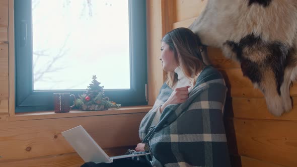Young Caucasian Woman Sitting Near the Window Covered with Blanket Lookin Outside and Using Laptop alt