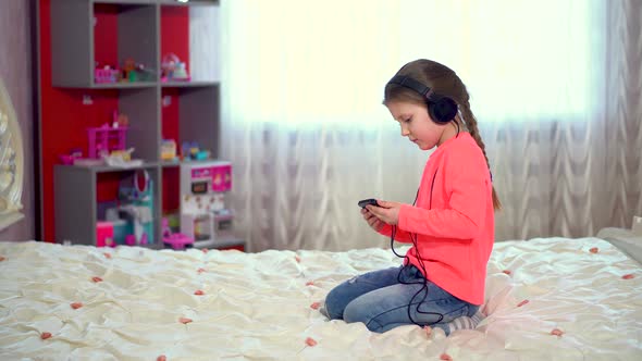 Beautiful little girl listening to music with headphones sitting on the bed in the bedroom