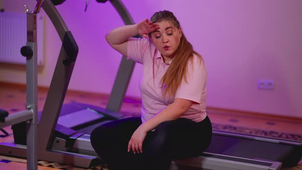 Portrait of Satisfied Young Chubby Woman Sitting on Treadmill Wiping Forehead with Hand alt