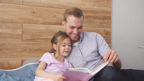 Family Father and Kid Girl Reading Book Fairytale Sitting on Bed at Home alt