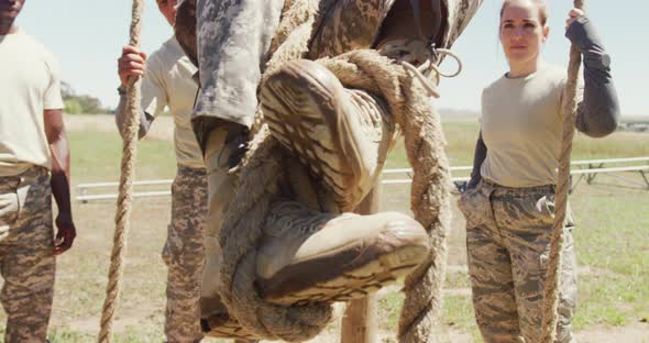 Diverse group watching male soldier in uniform climbing rope on army obstacle course in sun alt