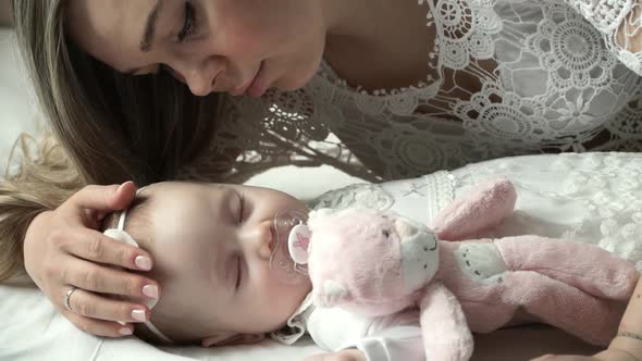 Close Up of Young Mother Kisses and Caresses Newborn Sleeping with Pacifier and Toy Baby Daughter