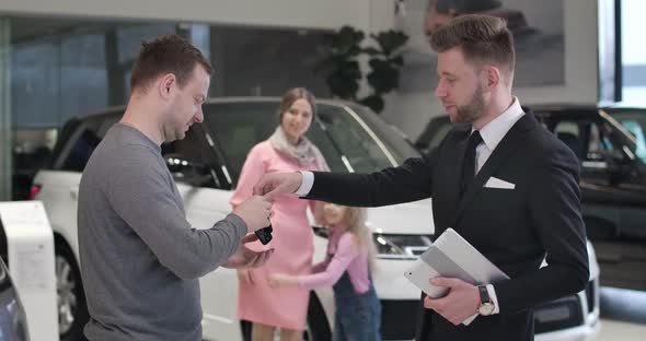 Young Caucasian Businessman Talking Car Keys and Shaking Trader's Hand As His Happy Wife alt