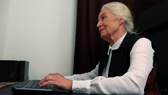 Old Happy Caucasian Woman Works on Computer in Home and Smiles alt