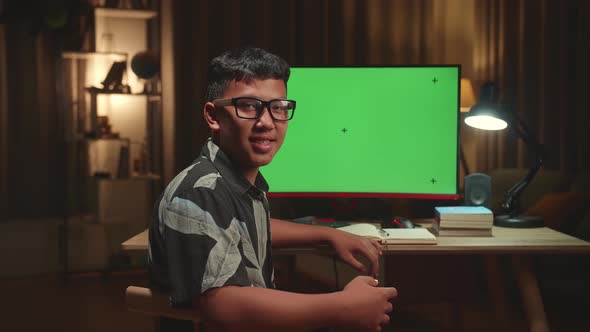 Asian Boy Learning Online With Computer Green Screen From Home, He Turns And Smiles Into The Camera alt