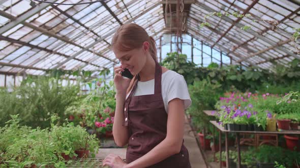 Young Girl Nursery Worker and Flower Greenhouse Speaks to a Client on the Phone
