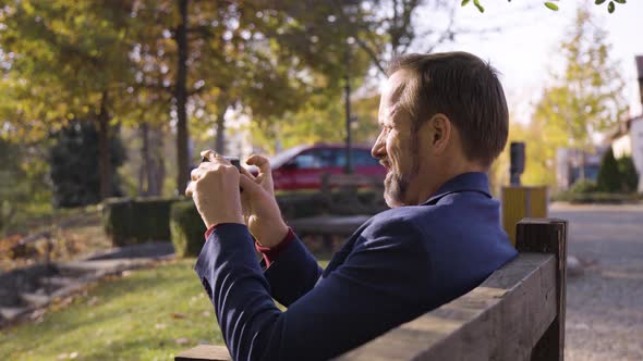 A Middleaged Handsome Caucasian Man Takes Pictures with a Smartphone As He Sits on a Bench