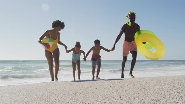 Smiling african american family with inflatables running on sunny beach alt