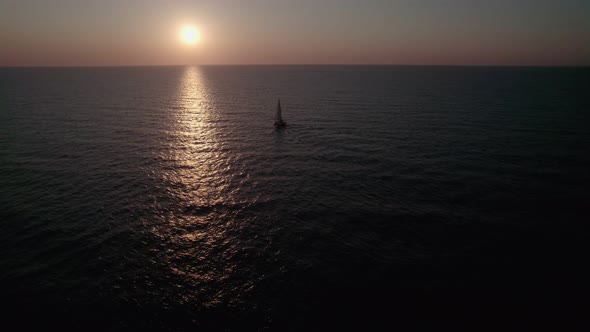 Sunrise over the sea, the camera flies over the water. A sailboat slowly sails against the sky.