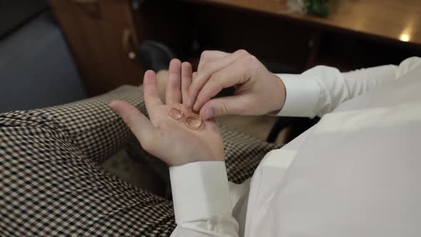 Groom in a White Shirt Holding Wedding Rings on the Palm of His Hand. Man Touches the Rings alt
