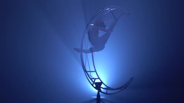 Gymnast Hangs Upside Down in a Twine on a Rotating Metal Construction . Blue Smoke Background alt