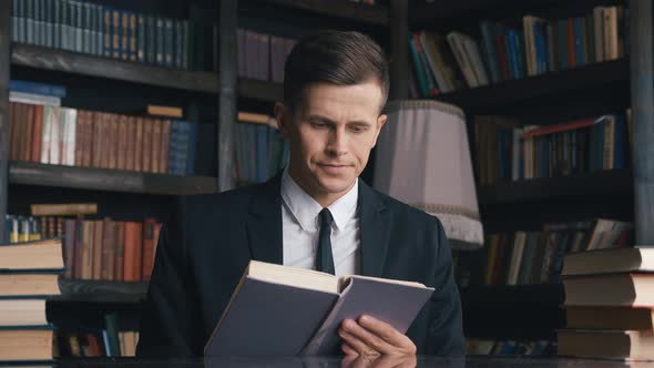 Handsome Businessman in Classical Suit Sitting in Library and Reading Book