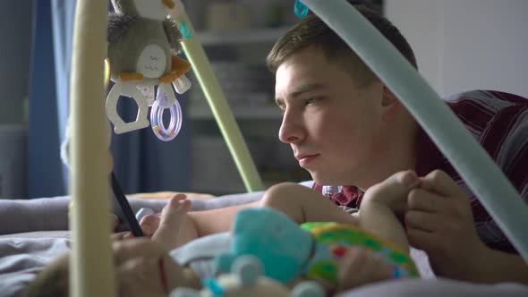 A Young Father Lies on the Bed with a Phone in His Hands Along with the Baby alt
