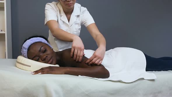 Female Masseur Making Massage for Young African Woman in Spa Salon alt