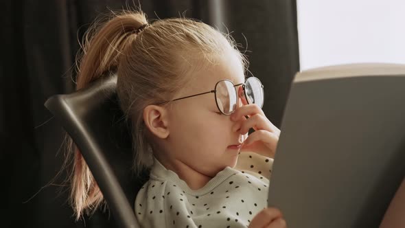 Little child tired girl reading a book in glasses after school rubs eyes from fatigue alt