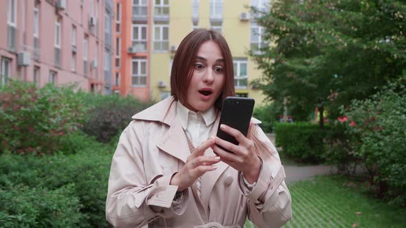 Girl Looks at the Phone and is Surprised at What She Saw Over Colorful Buildings Background alt