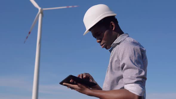 An AfricanAmerican Electrician in a Helmet Stands Against the Backdrop of a Windmill at an Air Power alt