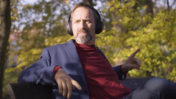 A Middleaged Caucasian Man Listens to Music with Headphones and Smiles As He Sits on a Bench