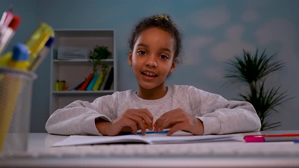 An African American Girl Sits at a Table in Front of a Laptop Talks on a Video Call Raises Her Hand alt