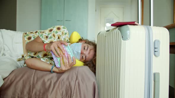 a Sad Tourist in a Mask Just From the Airport with a Suitcase is Lying on a Bad in a Hotel Room in alt