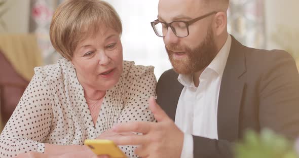 Handsome Businessman Teaching His Mature Grandmother Using Modern Smartphone. Good Looking Old Woman alt