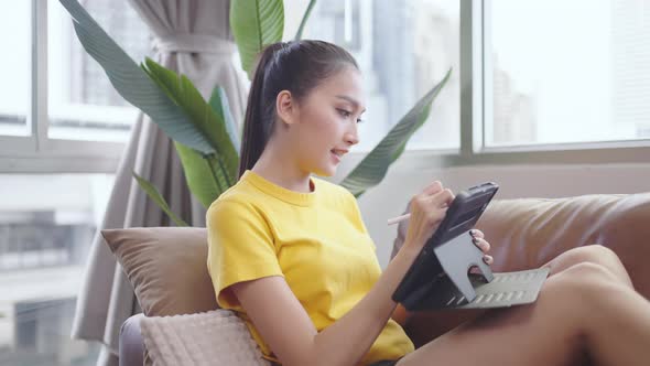 Young Asian woman using tablet and pen on sofa happy and smile.