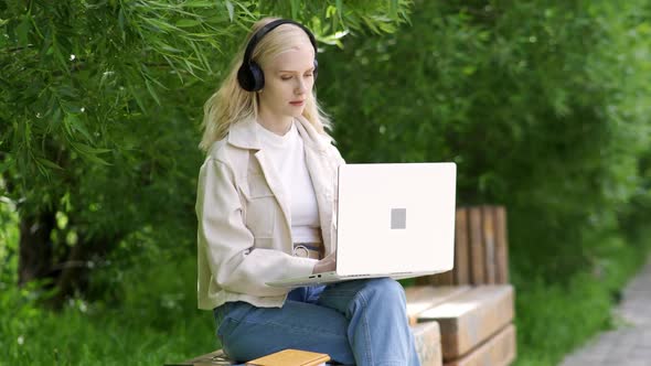 Young Beautiful Blonde Woman in Headphones Sits on a Park Bench with a Laptop alt