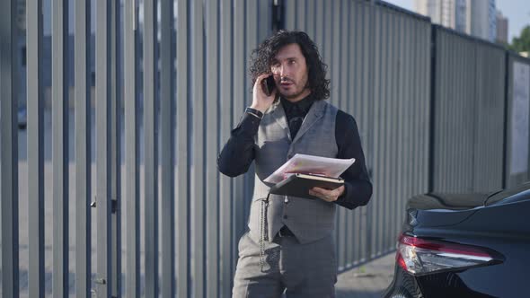 Busy Handsome Manager Standing in Urban City at Car with Paperwork Talking on Phone alt