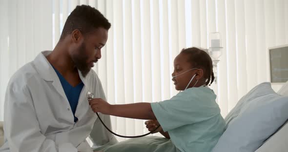 Cute African Child Playing Doctor with Man Pediatrician on Bed in Hospital Ward alt