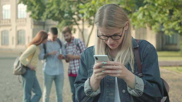 Upset Blond Girl Scrolling Smartphone Screen As Blurred Groupmates Laughing at the Background. Nerd alt