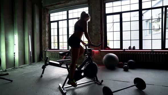 Young Sexy Sportswoman Is Riding Exercise Bike in Gym, Silhouette View of Slender Female Body alt