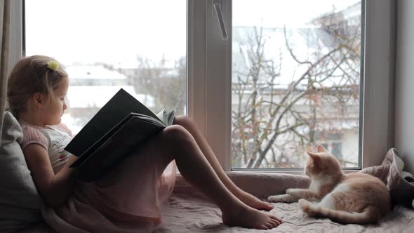 Little Girl Sits on a Windowsill with a Cat and Reads a Book