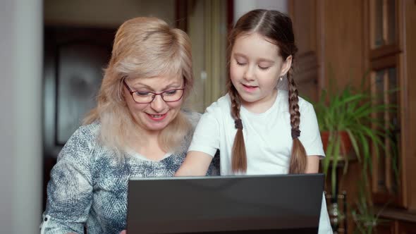 Happy middle aged grandma 55s and little preschool granddaughter playing games on laptop alt
