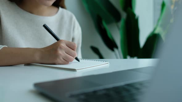 Closeup of a woman writing on a blank notebook with laptop computer on the table