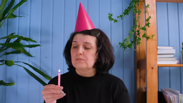 Upset Female Sitting Whit Bitrthday Hat and Looking with Sad Eyes on Camera Then Blowing Candle