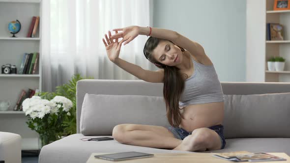 Future Mother Sitting on Couch in Headphones, Stretching Back, Audio Workout alt