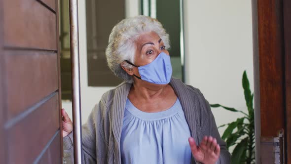 African american senior woman wearing face mask opening front door and welcoming at home alt