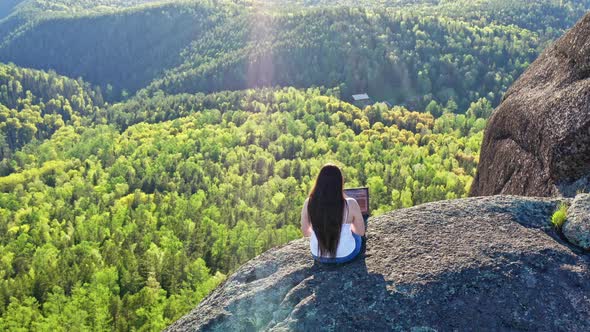 Aerial View of A Young Woman Working on a Laptop Sitting on a Mountaintop in the Forest