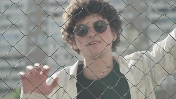 Close-up of Curly-haired Brunette Guy in Sunglasses Posing Behind Mesh Fence in Sunlight. Portrait alt