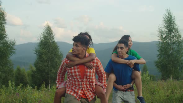 Happy Multiethnic Male Hikers Piggybacking Joyful Pretty Women on Mountain Trail at Sunset alt
