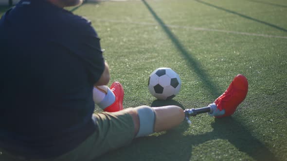 Zoom in to Soccer Ball on Green Grass with Unrecognizable Male Amputee Sitting on Stadium alt