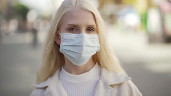 A Young Blueeyed Woman with Blond Hair in a Medical Mask Stands in the Middle of the Street