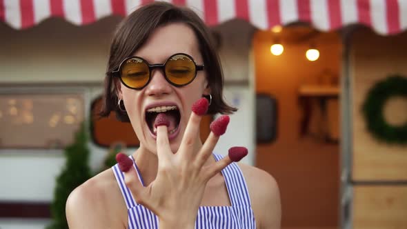 Stylish Caucasian Woman with Short Hair and Sunglasses Eats Raspberry From Fingers alt