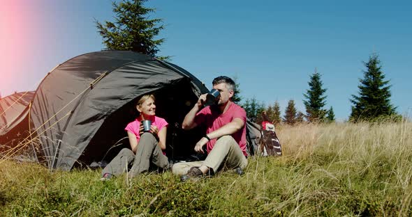 Young Man and Woman Holding Cups with Drinks Sitting Near the Tent and the Car in the Mountain alt