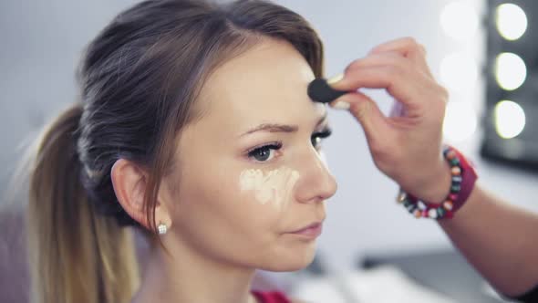 Closeup of Professional Makeup Artist Applying Concealer to Skin of Young Woman with Brush to Cover alt