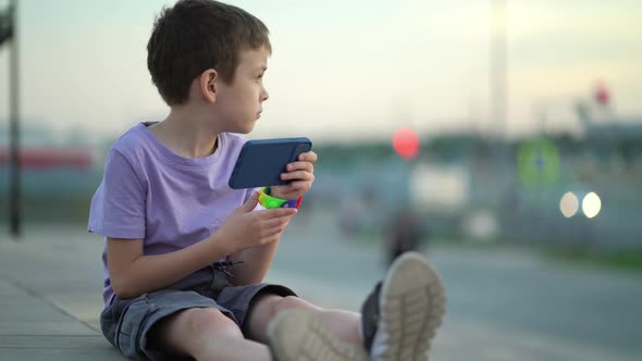 A Thoughtful Child Sits on the Steps on the Sidewalk and Looks Around and Into the Phone alt