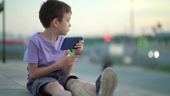 A Thoughtful Child Sits on the Steps on the Sidewalk and Looks Around and Into the Phone