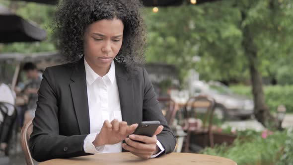 African Woman Cheering Success on Smartphone Sitting in Outdoor Cafe alt